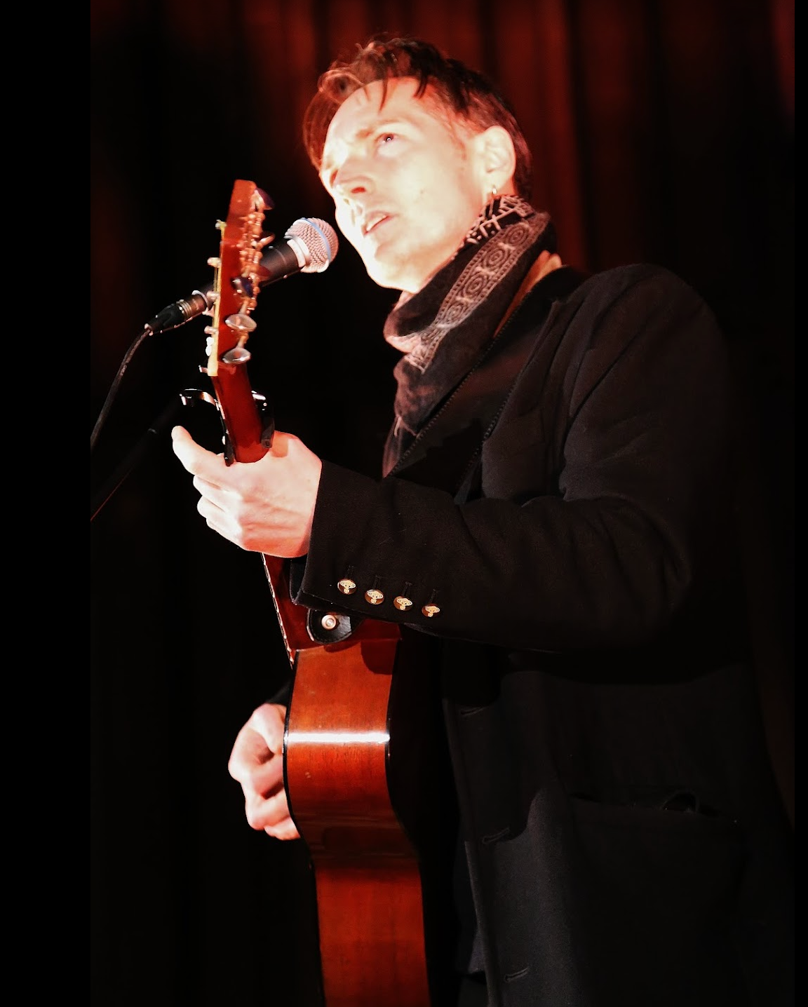 Man singing and playing acoustic guitar on stage under purple lights.