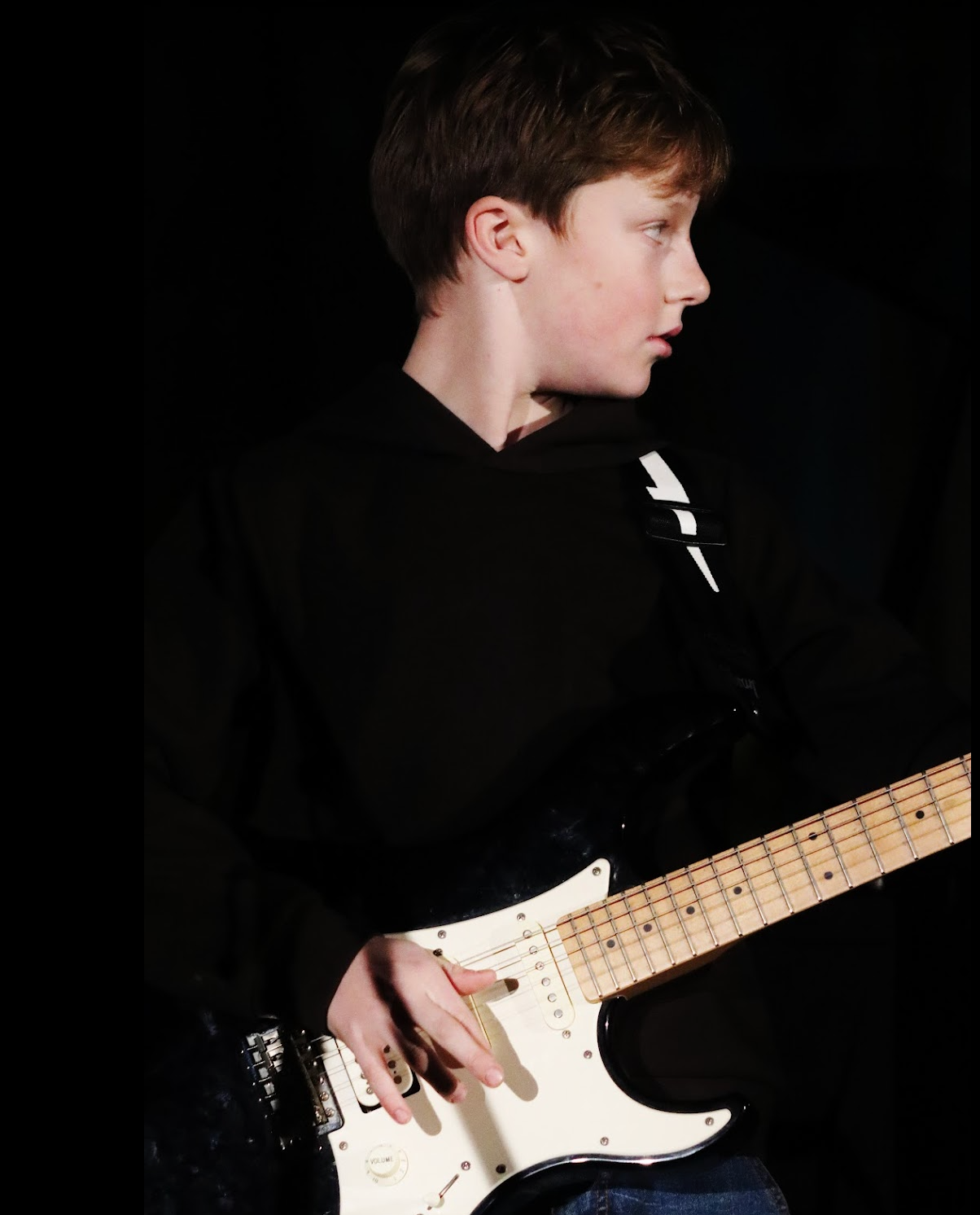 Young person playing guitar and singing on stage. Dark blue top, gray pants, light skin.