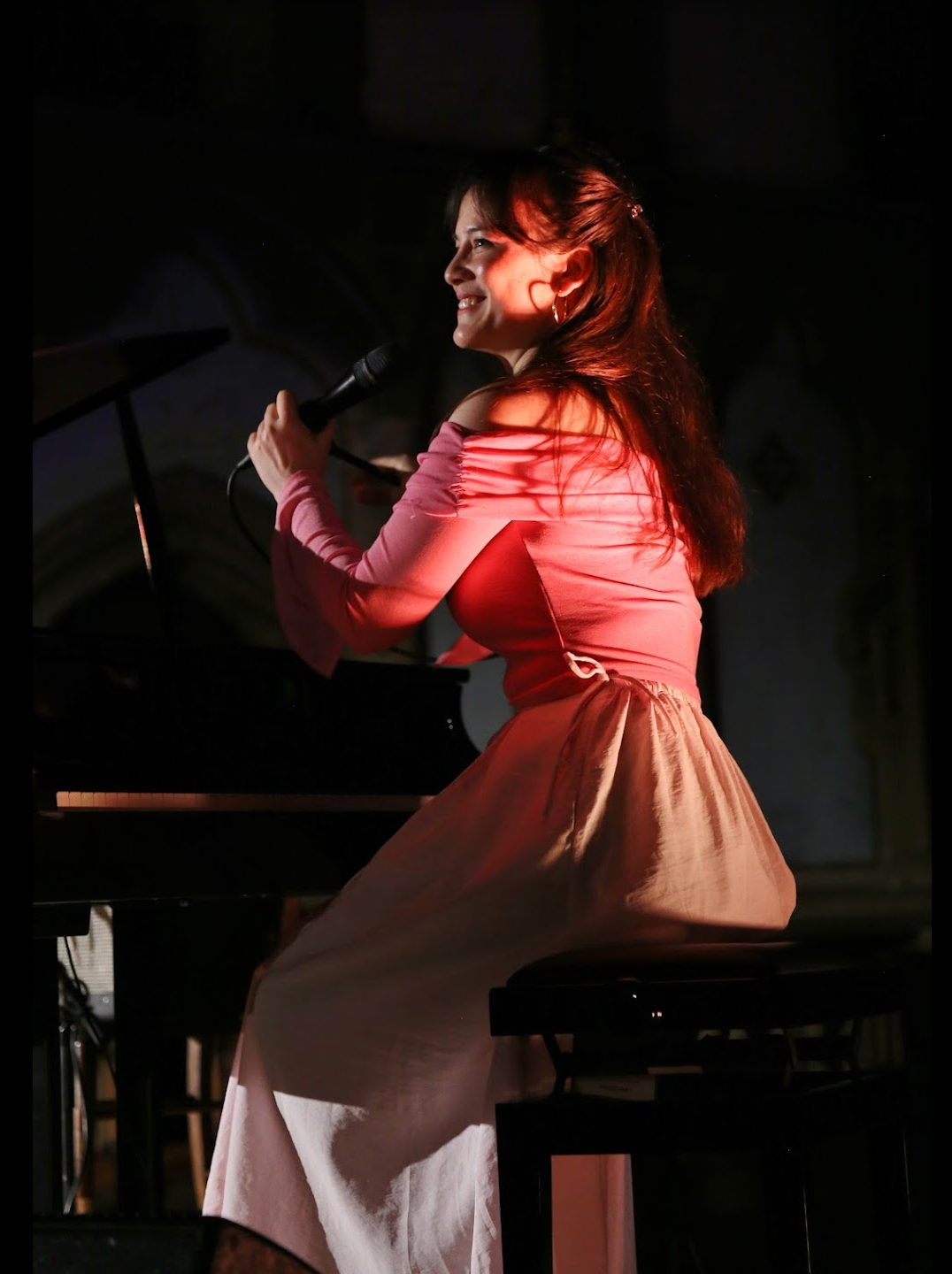 Woman singing and playing piano on stage, wearing pink top and white skirt.