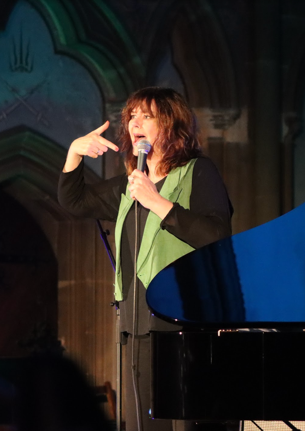 Woman speaking into a microphone, wearing black and green, standing on a stage. Man stands behind her.