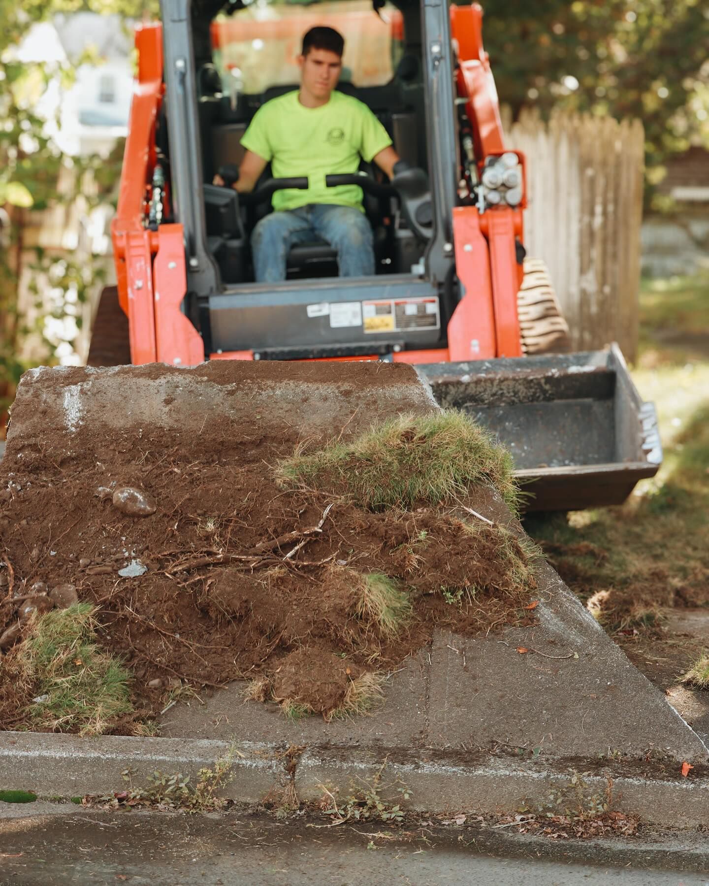 Man operating a Kubota skid steer, scooping up dirt and grass on a street.