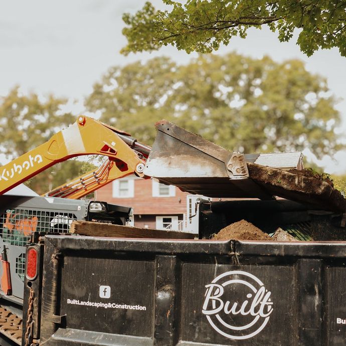 Orange excavator loading dirt into a black dump truck; red brick house in the background.