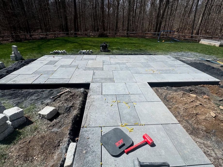 Patio construction: gray stone pavers being laid with a red mallet and tools on dirt and grass.
