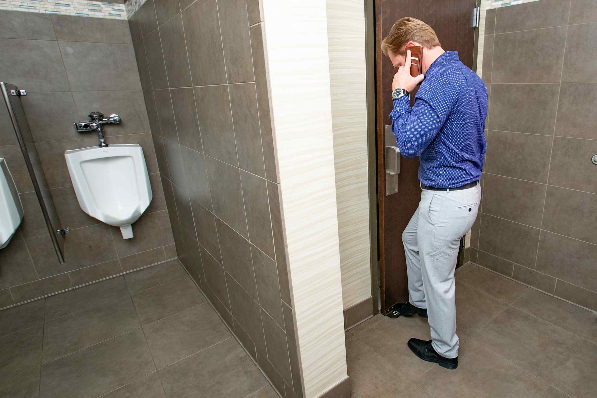 A man is standing in a public restroom talking on a cell phone.