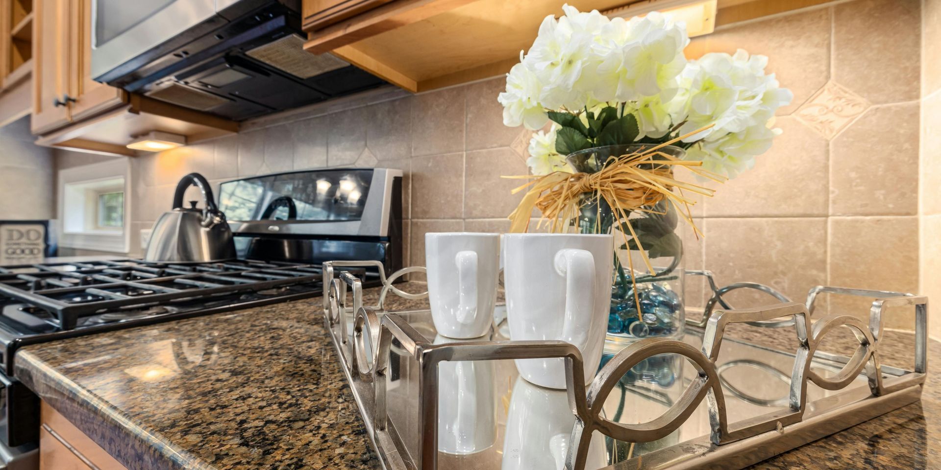 A kitchen with a decorative tray holding white mugs and flowers, in front of a stove.