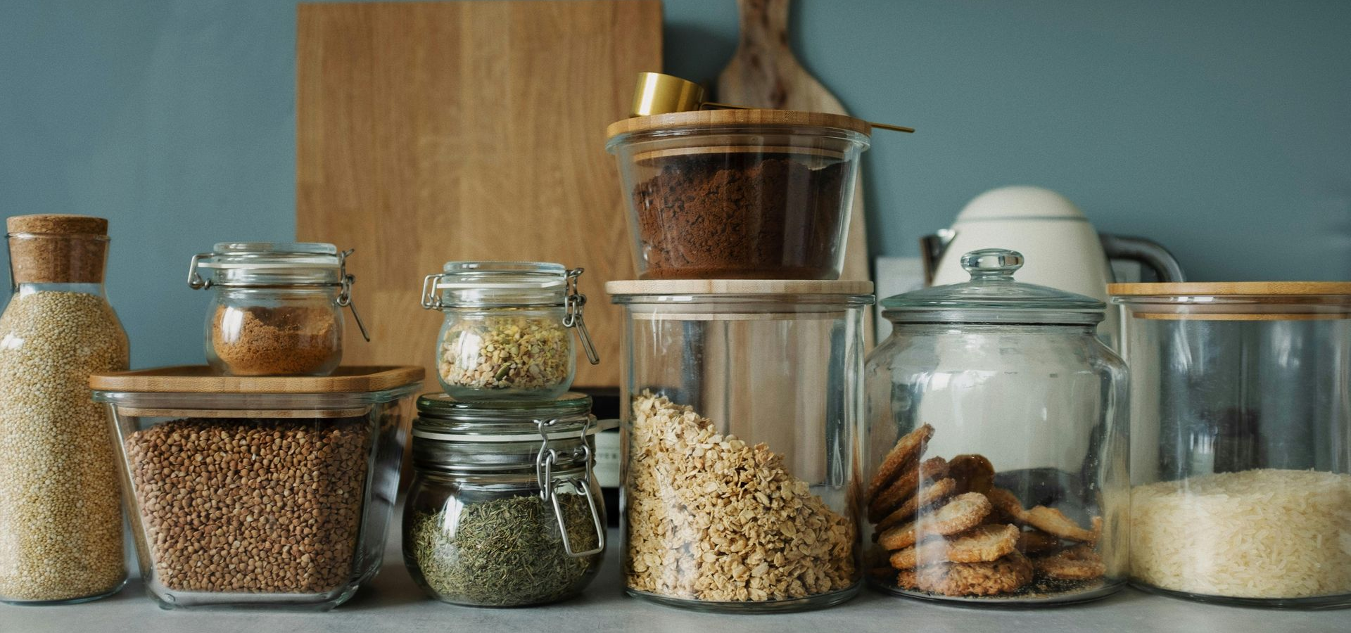 Glass jars filled with various ingredients are arranged on a shelf against a blue wall and wooden cutting board.