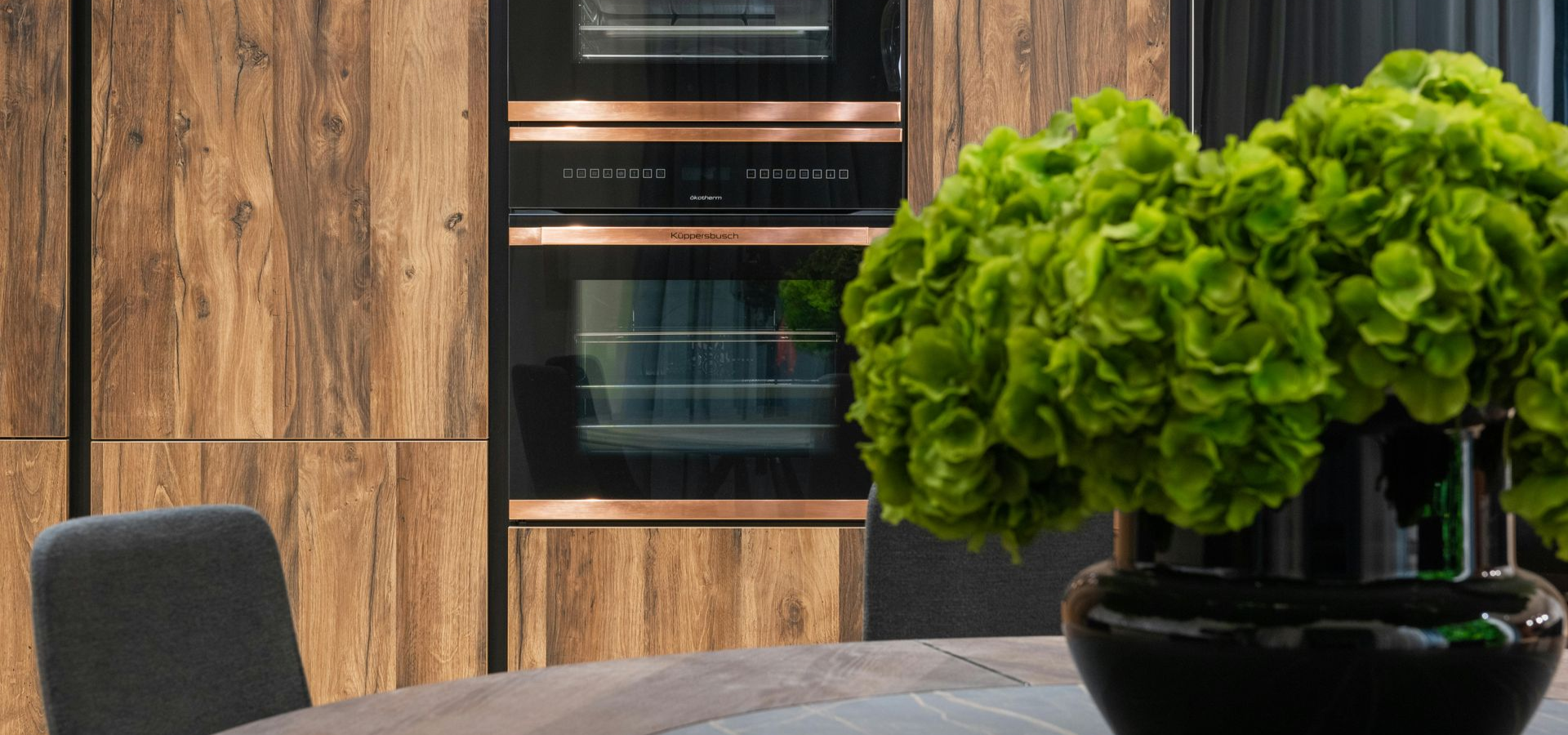 A kitchen with wood cabinets, black oven, and a large green floral arrangement in a black vase on a table.