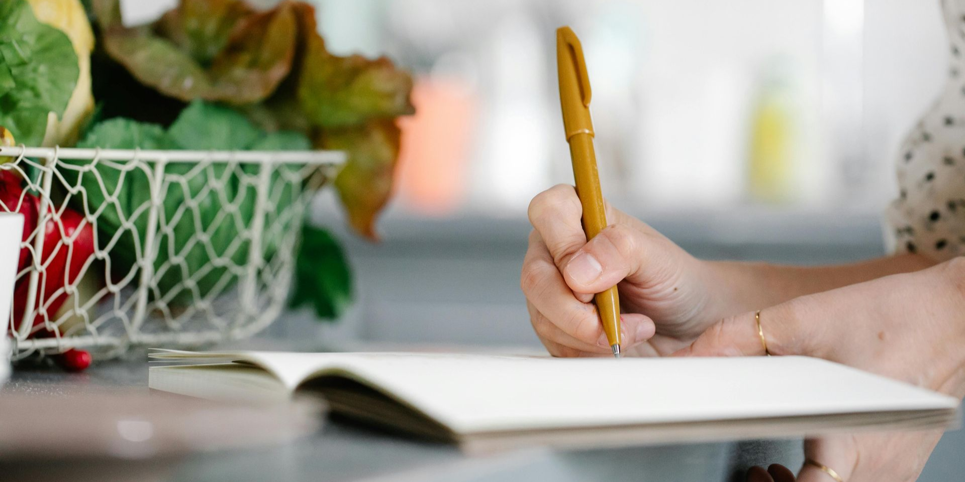A person writing in a notebook with a gold pen, beside a basket of green plants.