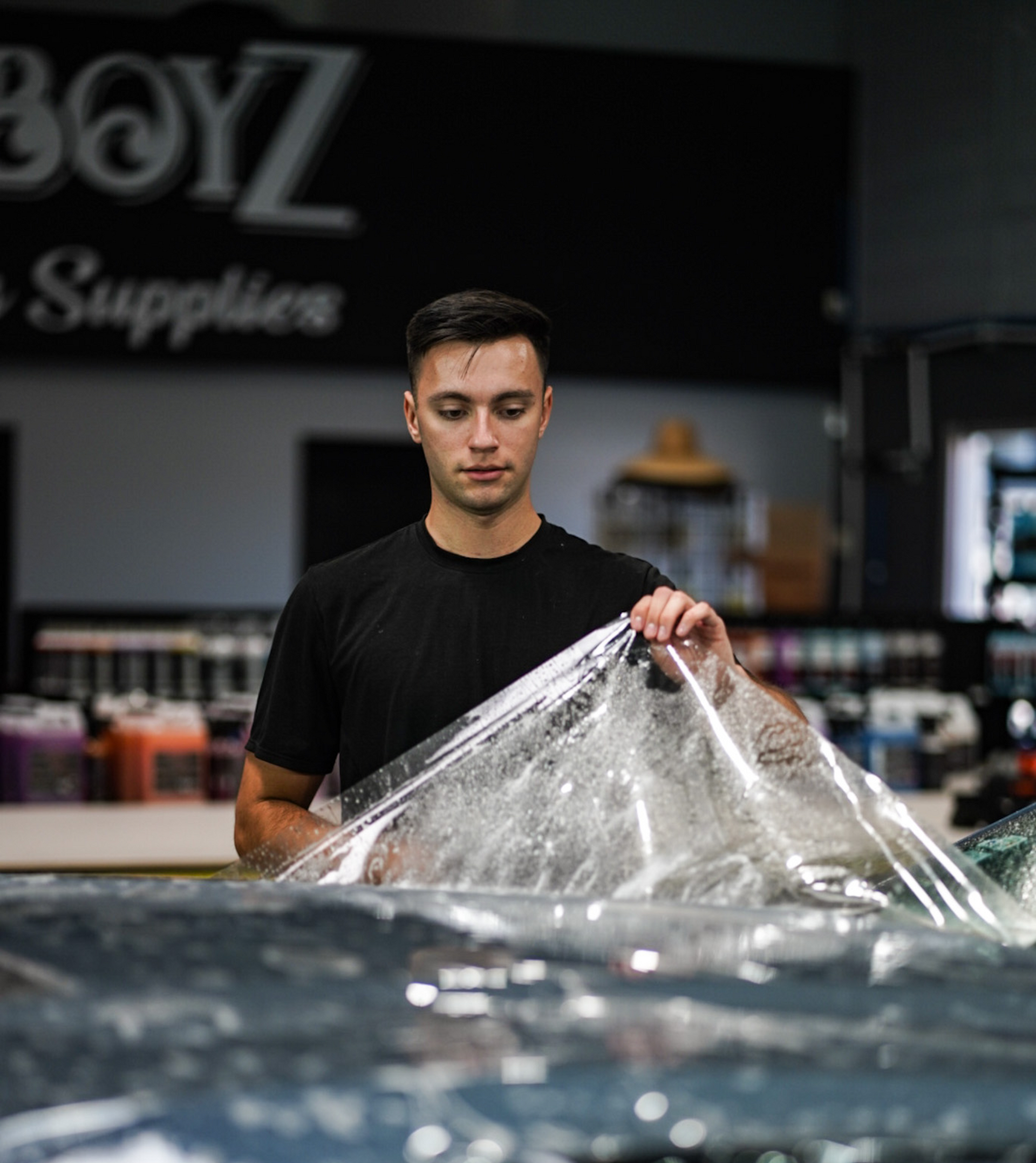 Man applying clear film to a car's hood in a shop. He is focused, using both hands.