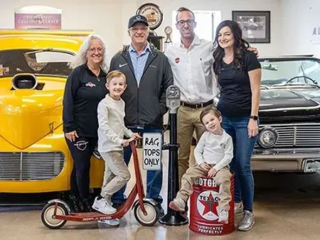 Family poses with vintage cars, a boy on a scooter, another on a gas can, in a garage.