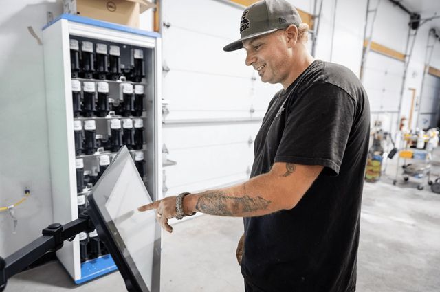 Man in cap smiles, pointing at touchscreen monitor next to a cabinet with black objects. Industrial setting.