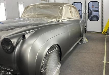 Classic silver car in a paint booth, covered in plastic for painting.