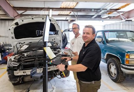 Two men in a garage, one using a laptop to assess a car with front-end damage; a green truck is in the background.