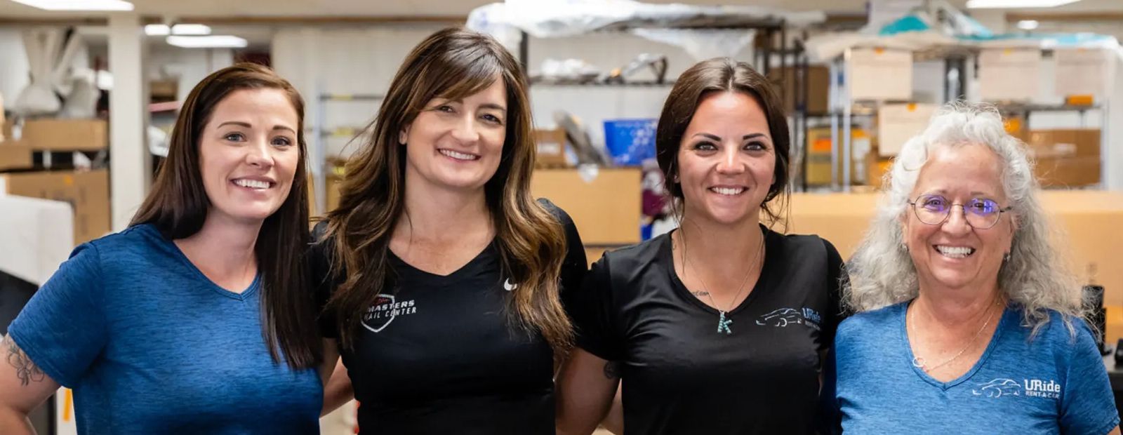 Four smiling women standing together in a workshop setting, wearing blue and black t-shirts.