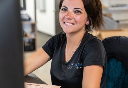 Woman smiling at a desk, wearing a black shirt with a logo.