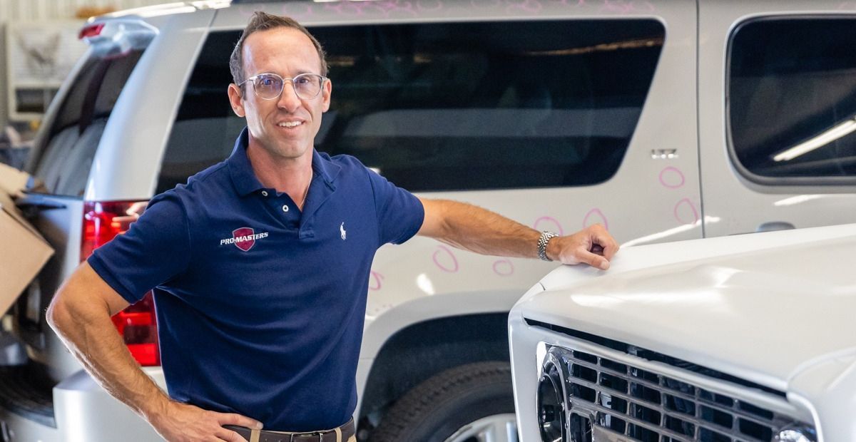 Man in blue polo shirt leaning on white SUV in a garage, smiling. Another SUV in the background.