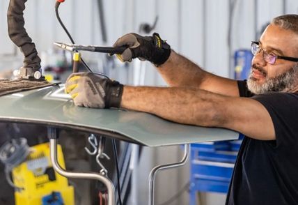 Auto body worker uses a tool to repair a dent on a car's hood in a workshop.