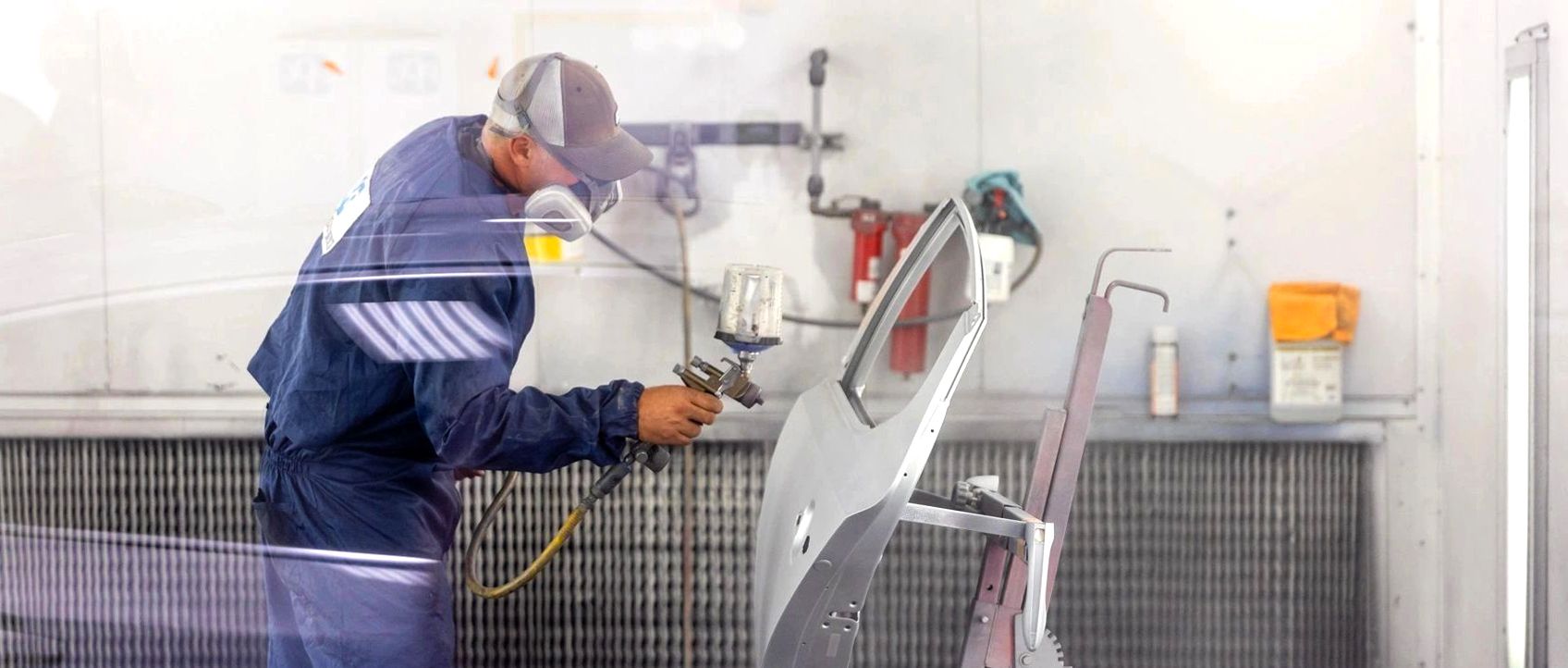 Person in a paint booth spraying a car door; wearing a respirator, hat and blue work suit.