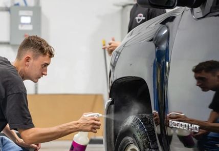 A person sprays a black truck's tire with a spray bottle in a shop. Another person is visible in the background.