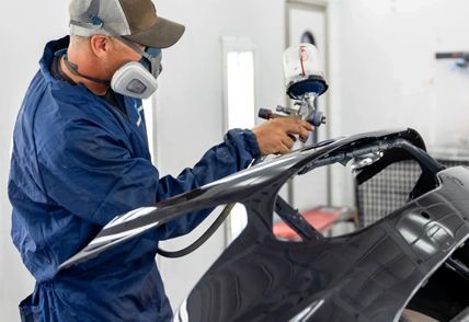 Person in protective gear sprays paint on car part in a workshop.