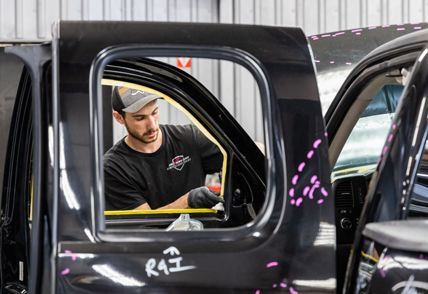 Person working on a black truck door inside a shop. He uses tools, wearing a hat, gloves.