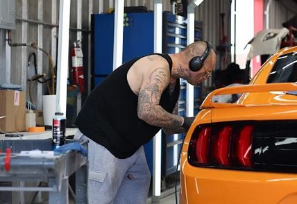 Man in a garage working on an orange sports car, wearing headphones.
