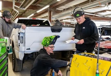 Three mechanics working on a white truck in a shop, one welding, two others observing and reviewing papers.