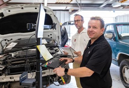 Two men in a car repair shop examining a car's engine. One types on a keyboard, the other holds a tablet.