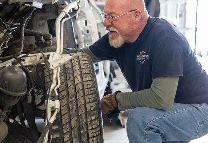 Mechanic examines a car tire. He wears a t-shirt, gloves, and glasses. White vehicle body in background.