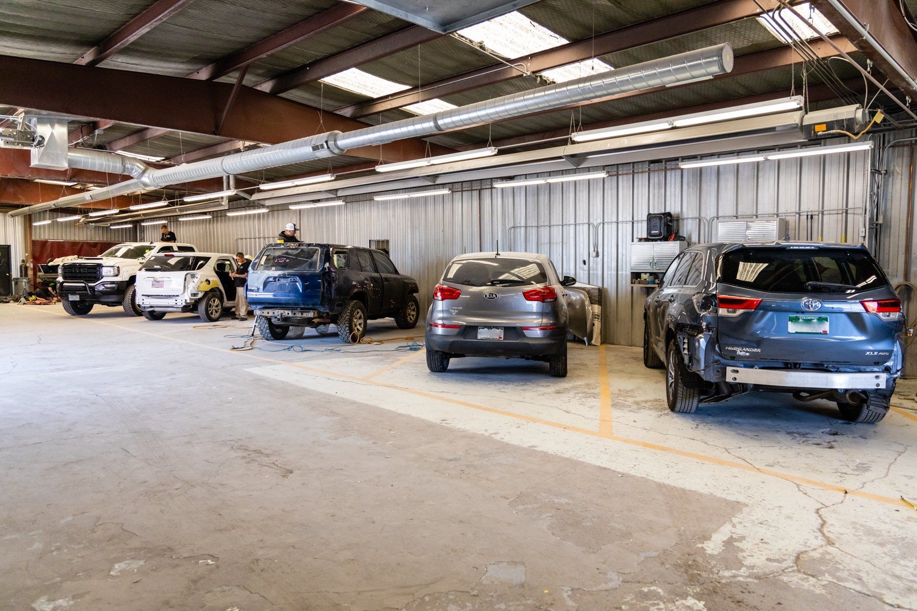 Cars in a repair shop; gray floor, white walls, overhead lights, damaged vehicles undergoing repairs.