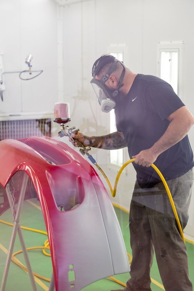 Person in mask sprays a red car bumper in a paint booth.