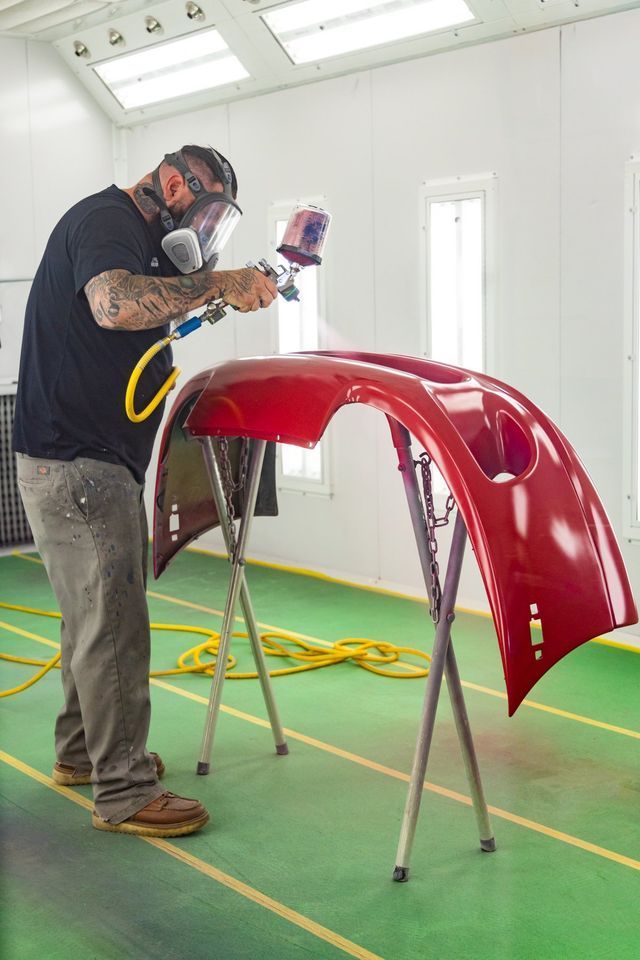 Man in respirator sprays red car bumper in a paint booth.