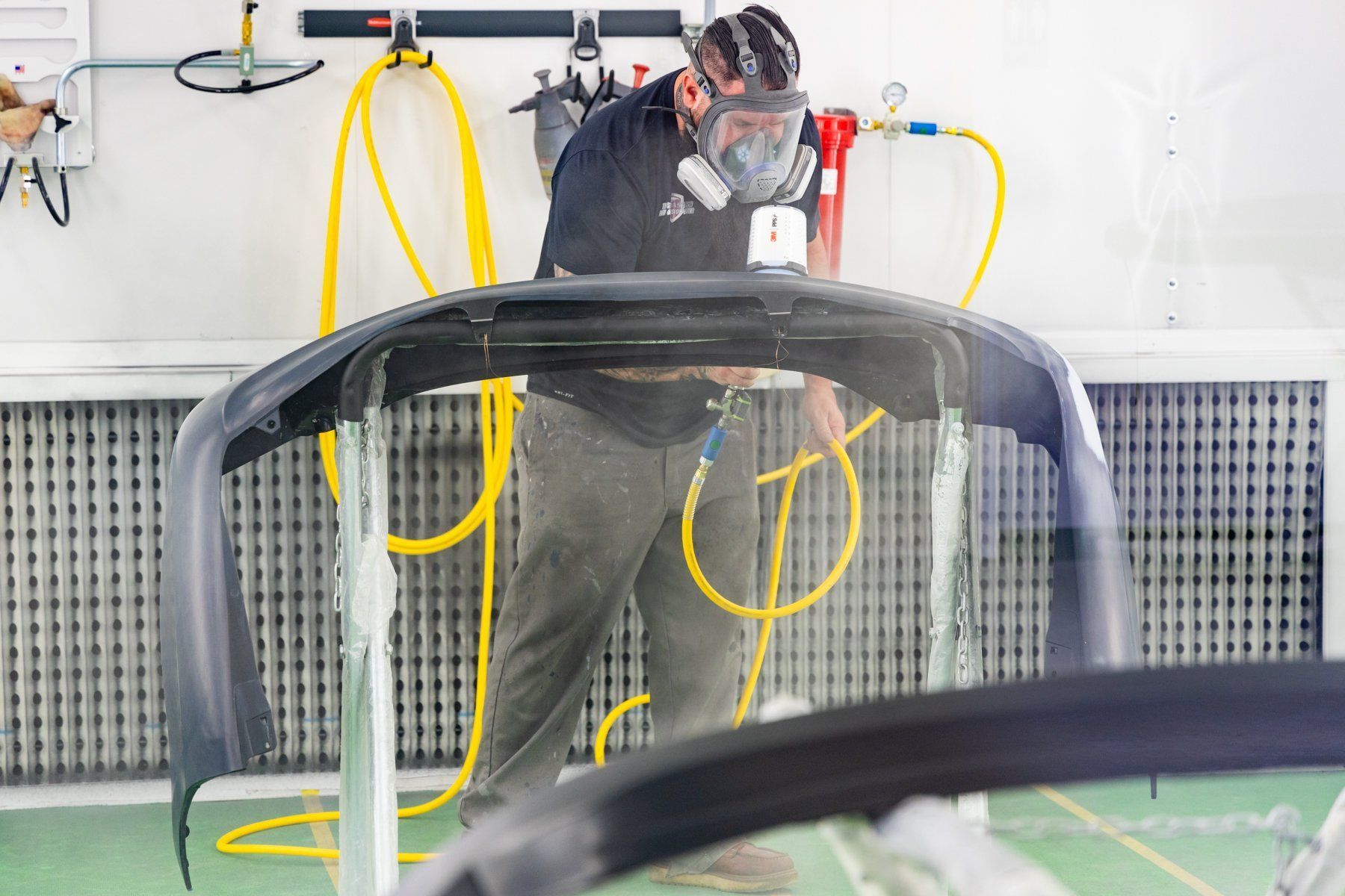 Man in respirator sprays a car bumper in a workshop, surrounded by yellow hoses and equipment.