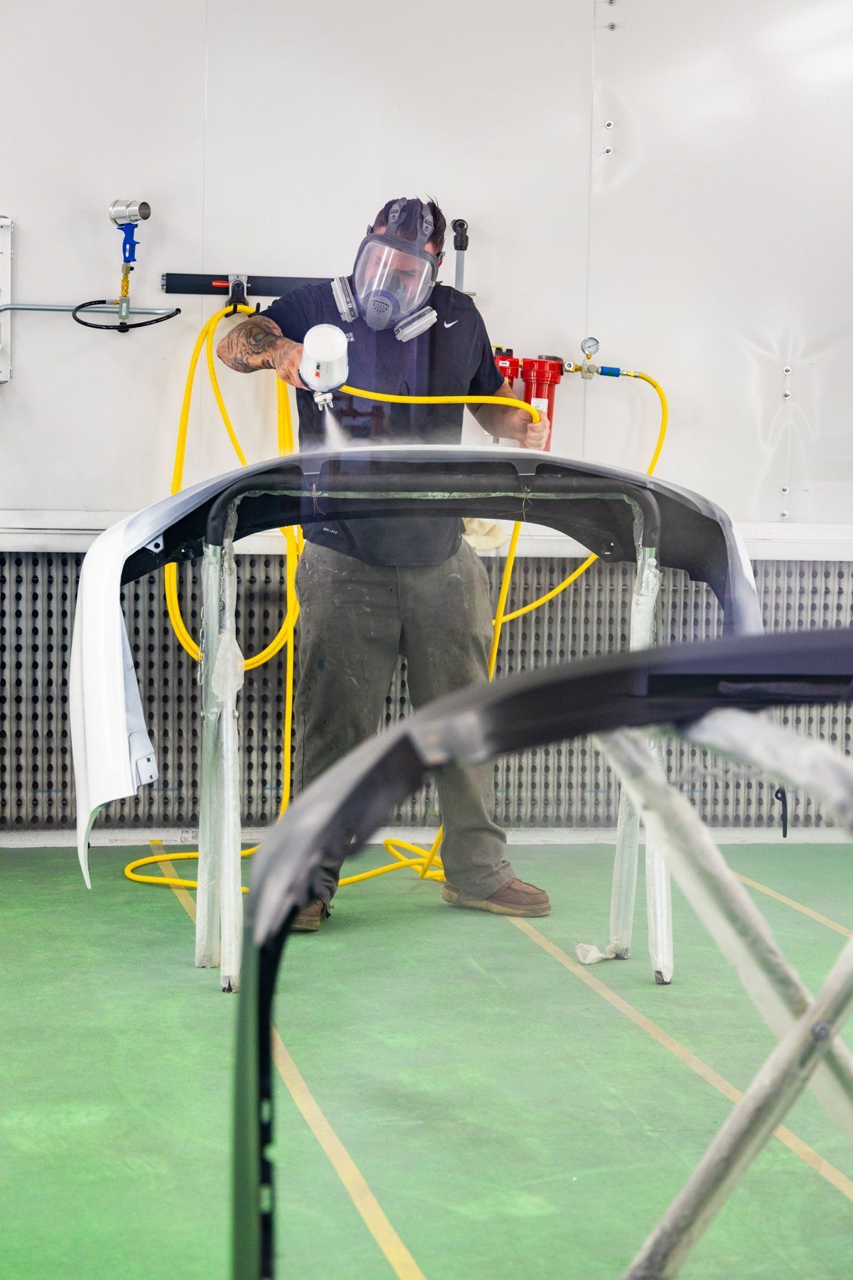 Person spraying a car bumper in a paint booth, wearing a respirator.