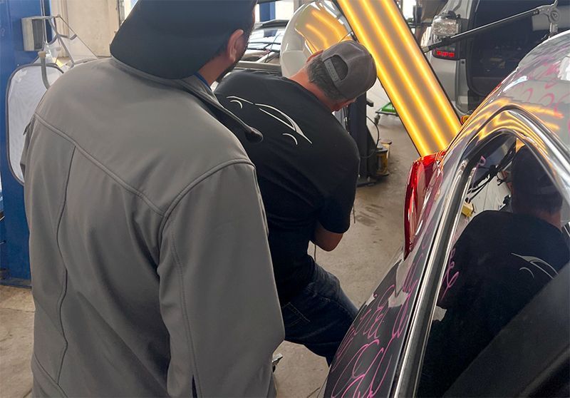Two men working on a car in a repair shop. One examines the vehicle's side, possibly dent removal.