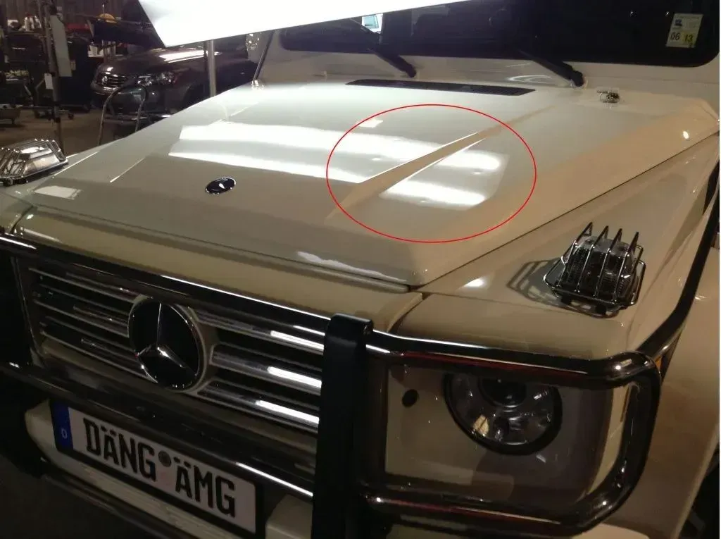 White Mercedes-Benz G-Class hood with a dent, circled in red. Silver grill, black brush guard, and a license plate.