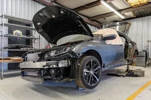Car in a repair shop with hood and door open, front damaged, surrounded by tools and equipment.