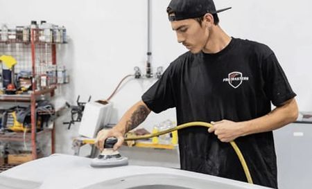 Person sanding a car panel in a workshop. Wearing black cap and shirt, holding sander and hose.