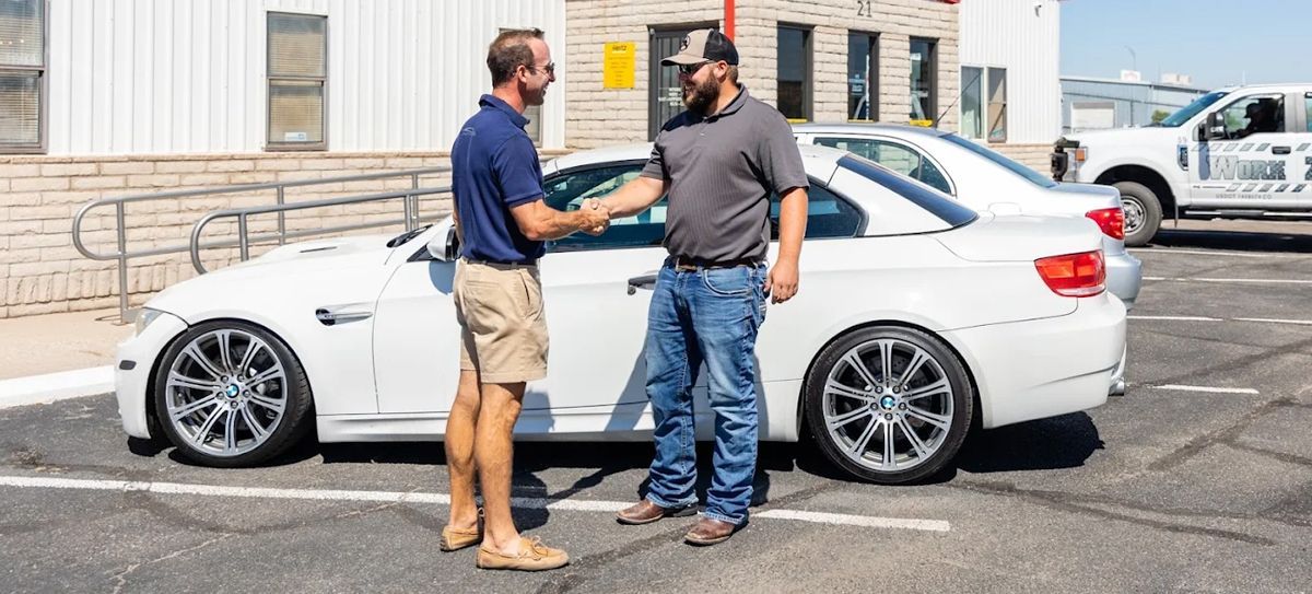Two men shaking hands beside a white car in a parking lot.