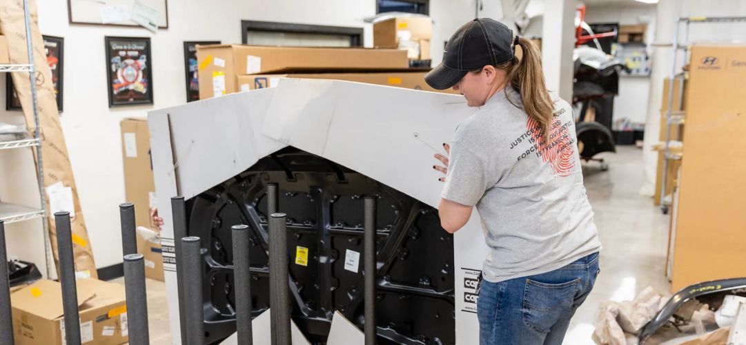 A person in a baseball cap carries a large packaged item in a warehouse.