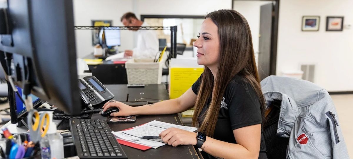 Woman working at a desk, typing on a computer. A man works in the background. Office setting with a jacket hanging.