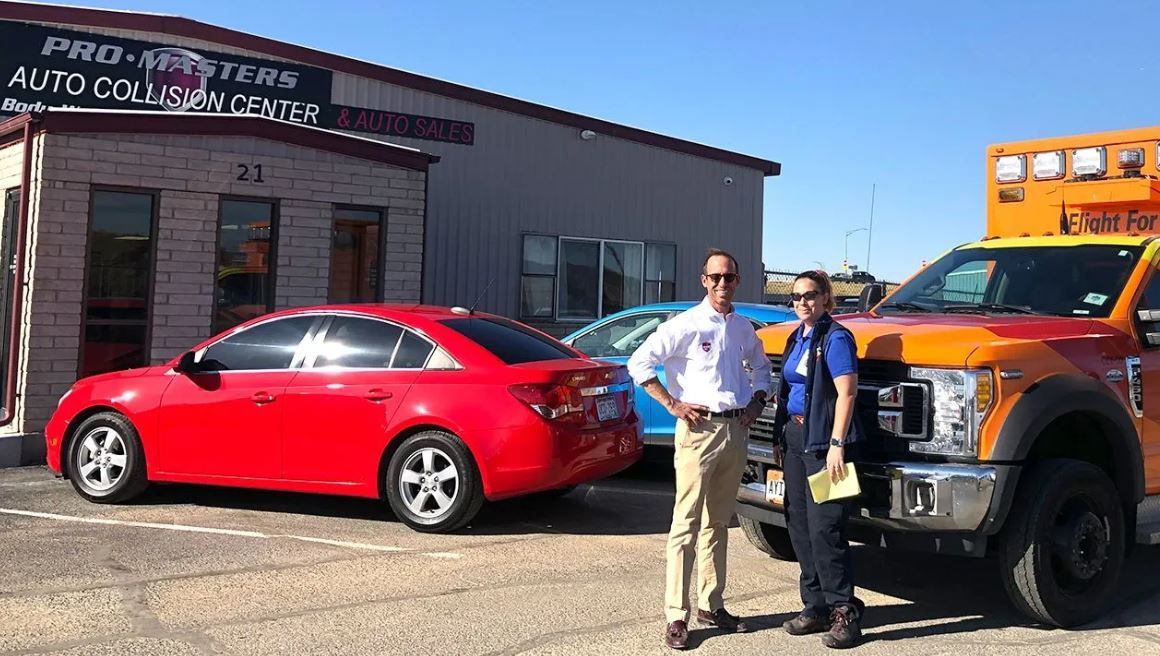 Two men stand near red car and orange truck at auto collision shop.