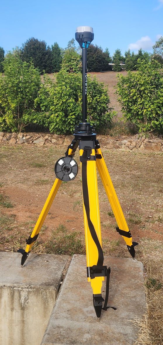 Surveying equipment on a yellow tripod in a field. The equipment is black and silver. Blue sky and green bushes in background.
