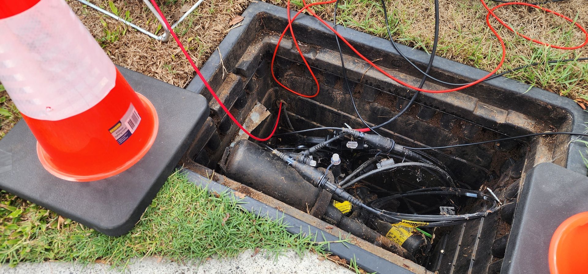 Open utility box with red wires, orange cone on a mat, grass background.