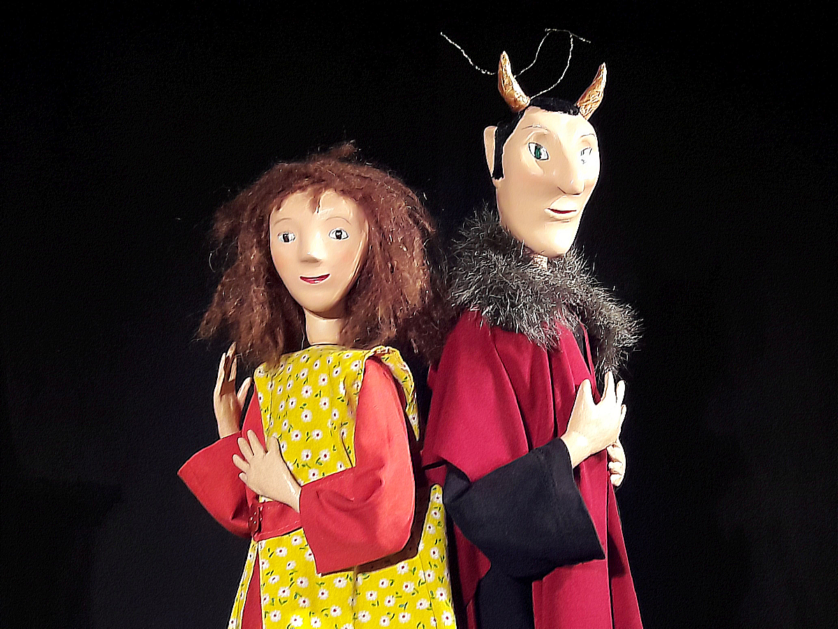 Zwei Handpuppen, eine mit wuscheligem braunem Haar in einem gelben Kleid und die andere mit kleinen Hörnern und einem roten Mantel, stehen vor schwarzem Hintergrund.