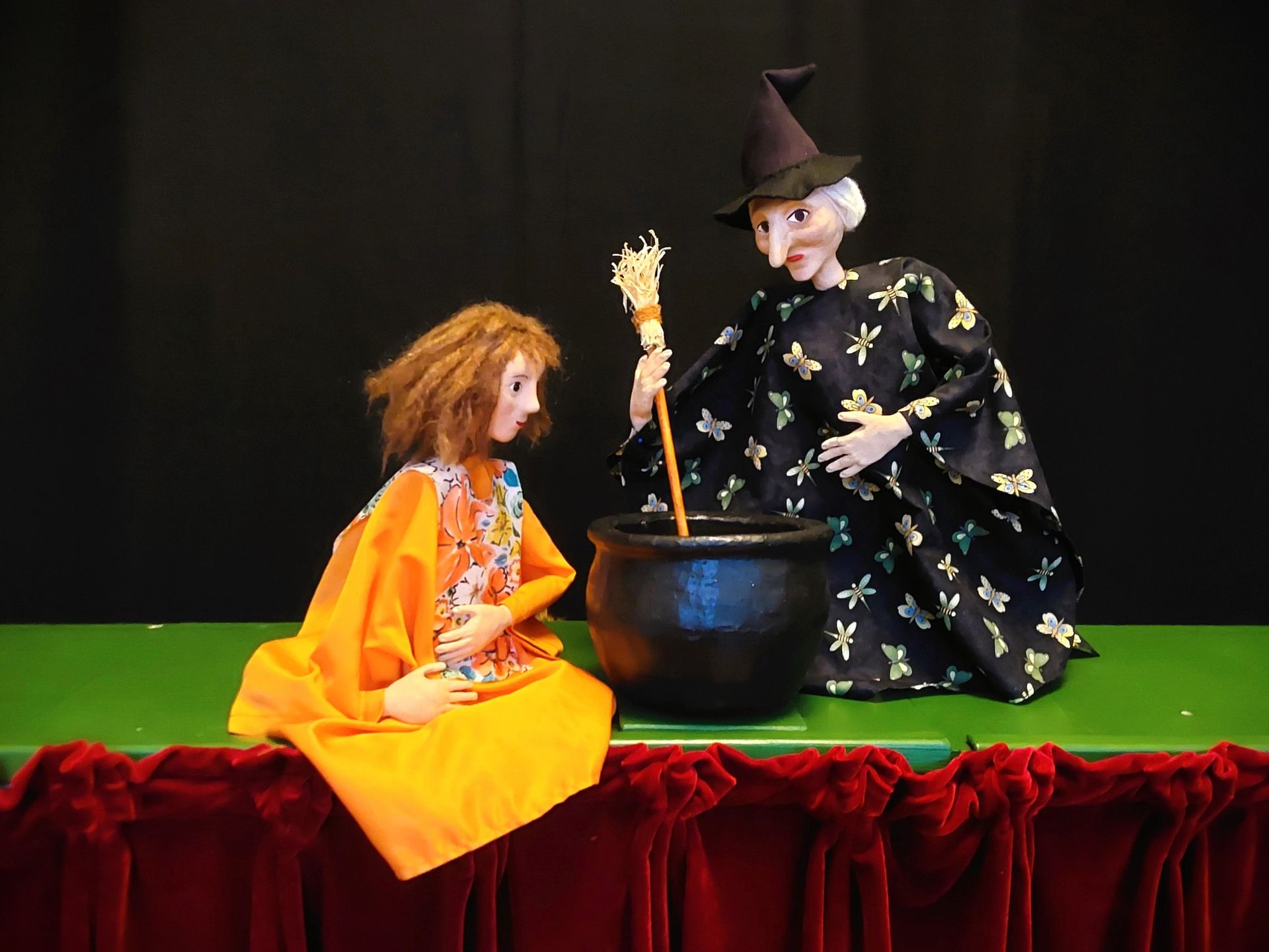 Zwei Handpuppen, eine mit wuscheligem braunem Haar in einem gelben Kleid und die andere mit kleinen Hörnern und einem roten Mantel, stehen vor schwarzem Hintergrund.