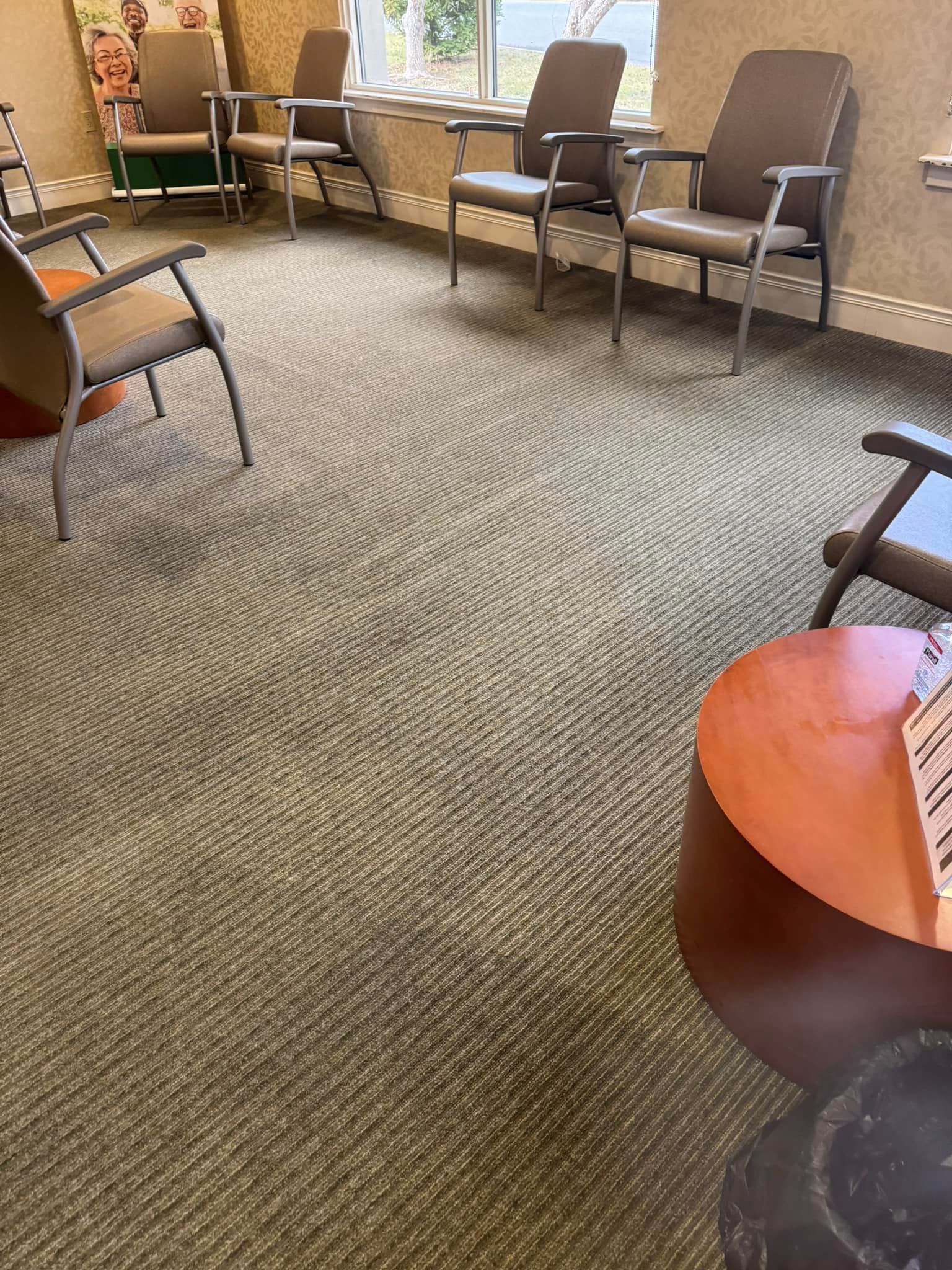 Waiting room with several chairs, circular table, and patterned carpet. Window in background.