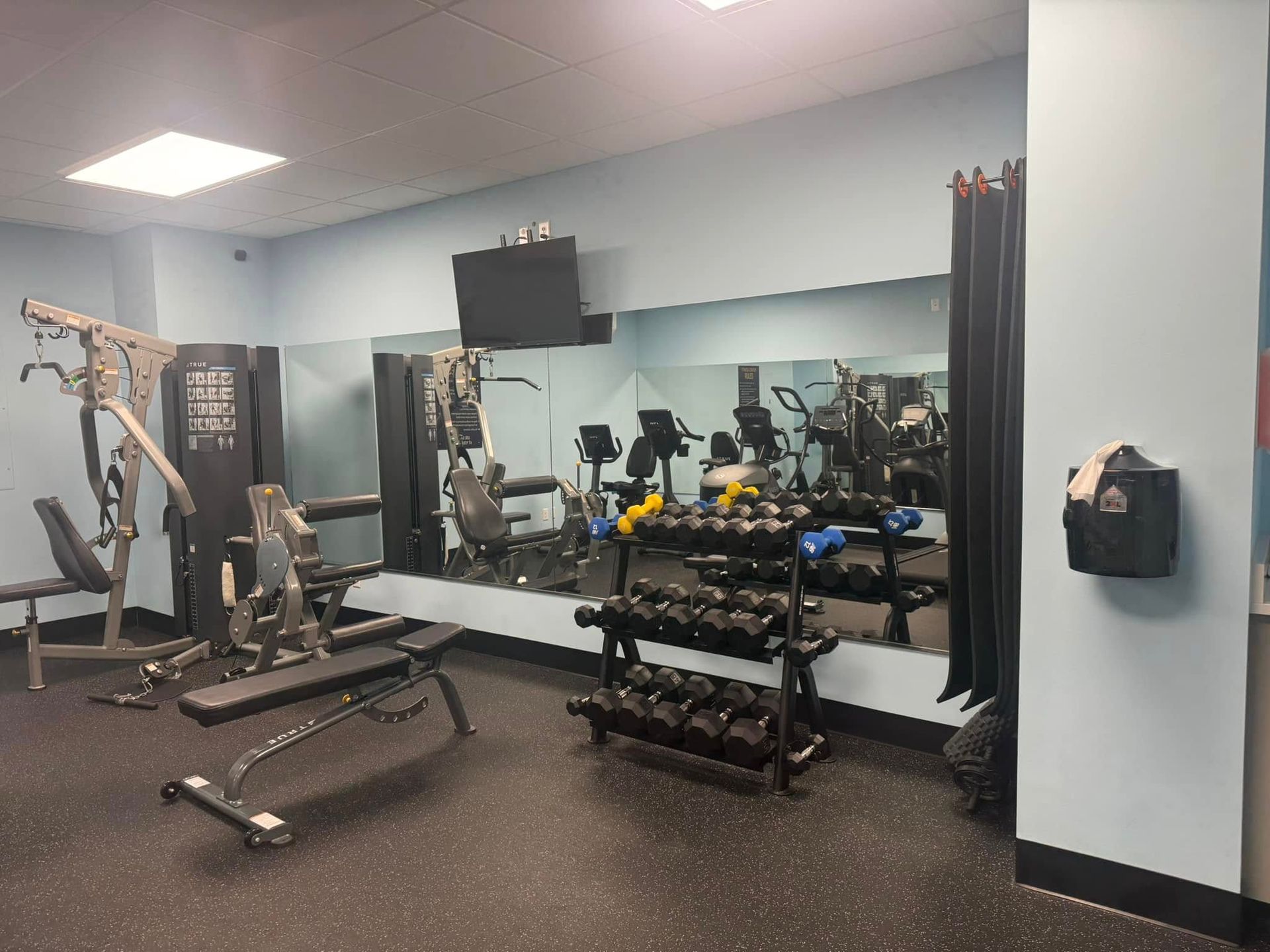 Gym with workout equipment, including weights, weight machines, and a bench. Reflective mirror on the wall. Gym with workout equipment, including weights, weight machines, and a bench. Reflective mirror on the wall.
