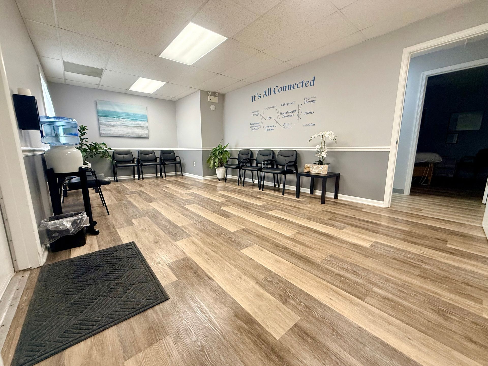 Waiting room with chairs, water dispenser, and artwork. Wooden floor, neutral walls.