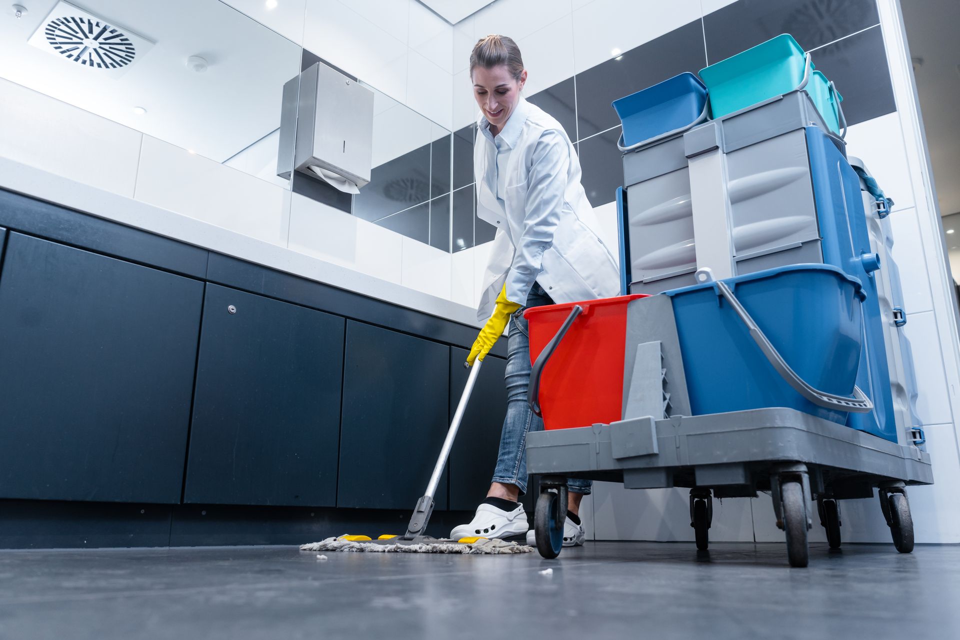 Person wearing gloves mops a floor in a modern bathroom, cleaning supplies cart nearby. Person wearing gloves mops a floor in a modern bathroom, cleaning supplies cart nearby.